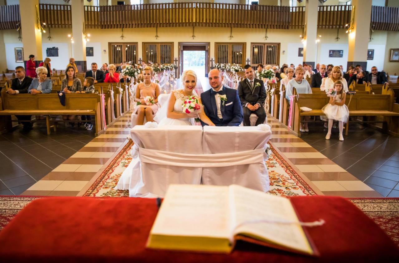 Para młoda siedzi przy ołtarzu podczas ceremonii ślubnej w kościele, goście siedzą w ławkach, widoczny otwarty śpiewnik na pierwszym planie.