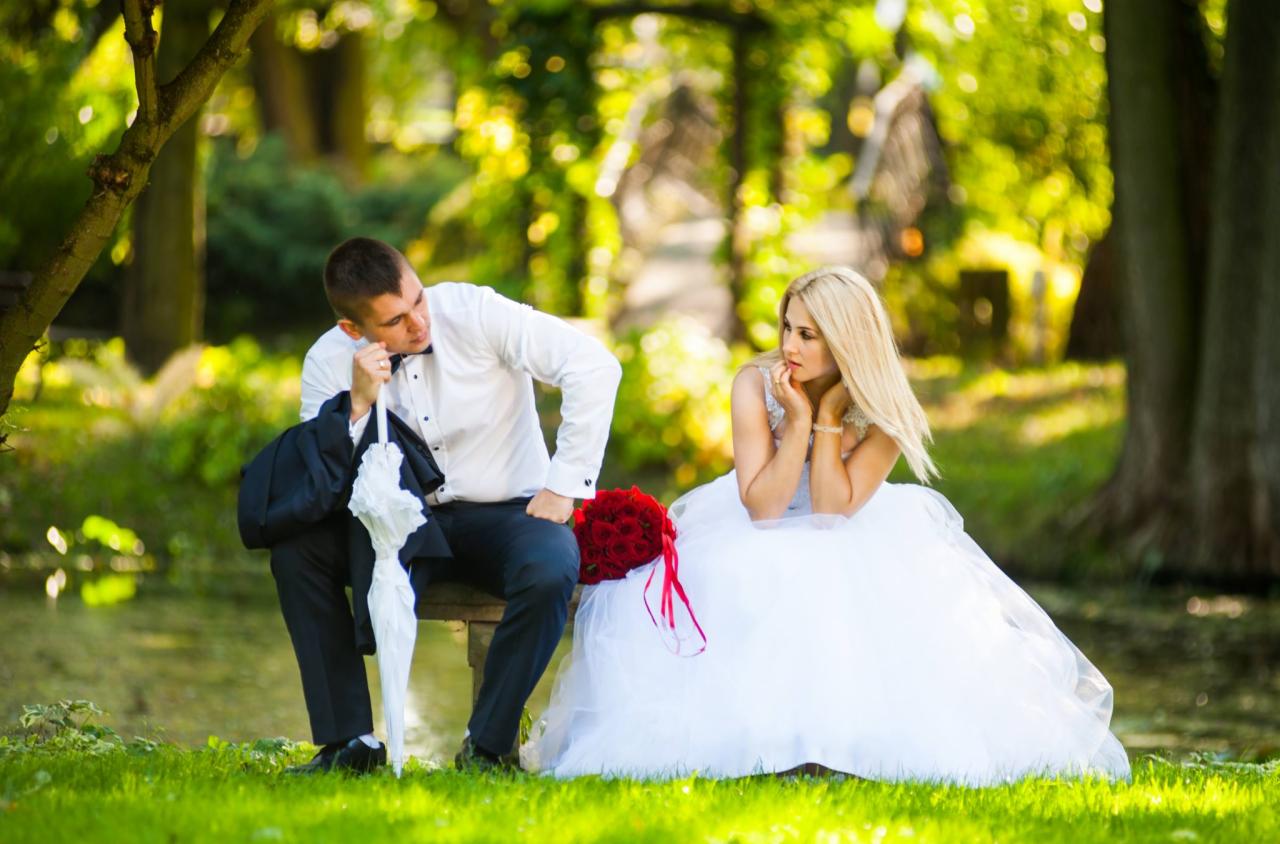 Zamyślona para młoda siedzi na ławce w parku obok stawu. Pan młody w białej koszuli i muszce opiera się o białą parasolkę, obok leży jego marynarka. Panna młoda w białej sukni z bukietem czerwonych...