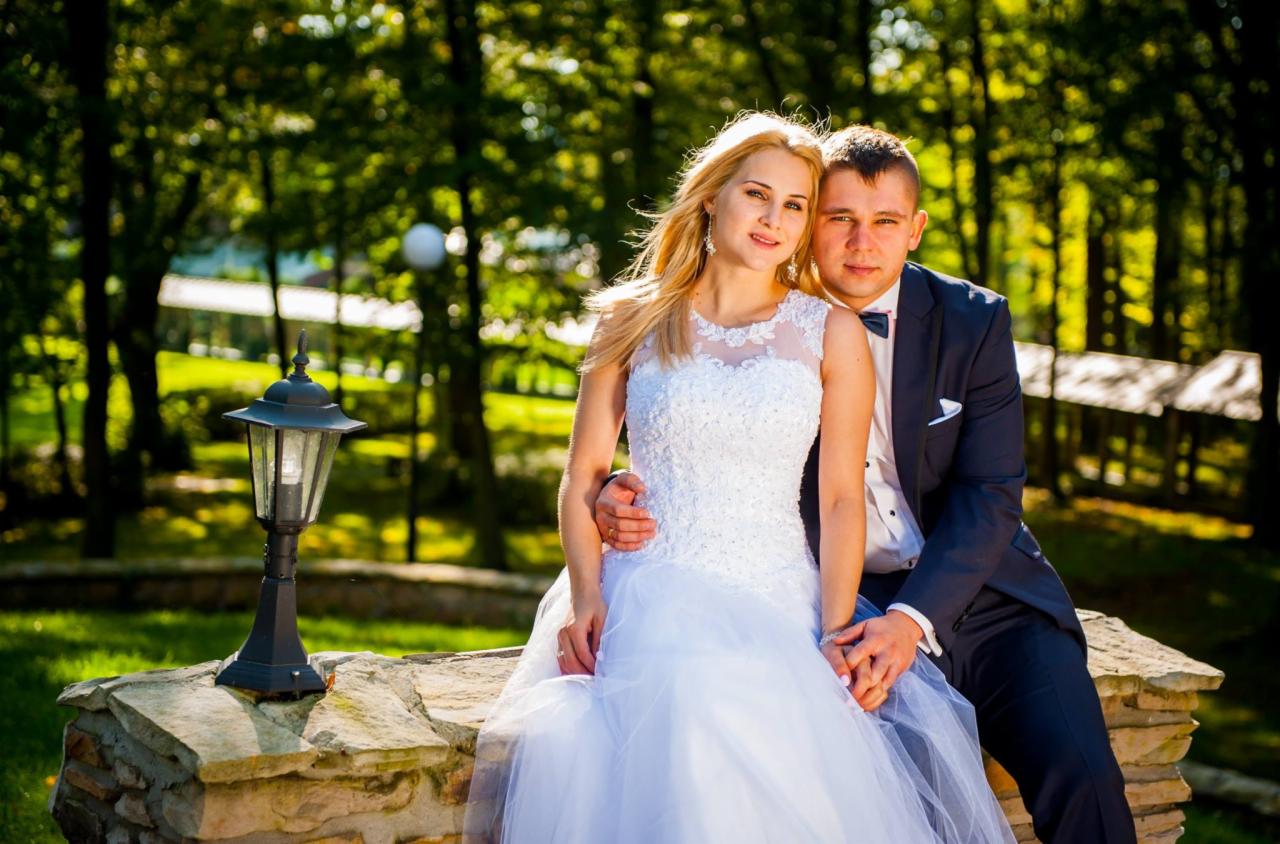 Para młoda siedząca na kamiennym murku w parku, panna młoda w białej sukni z koronkową górą i tiulowym dołem, pan młody w granatowym garniturze z muszką, w tle zielone drzewa i lampa parkowa.