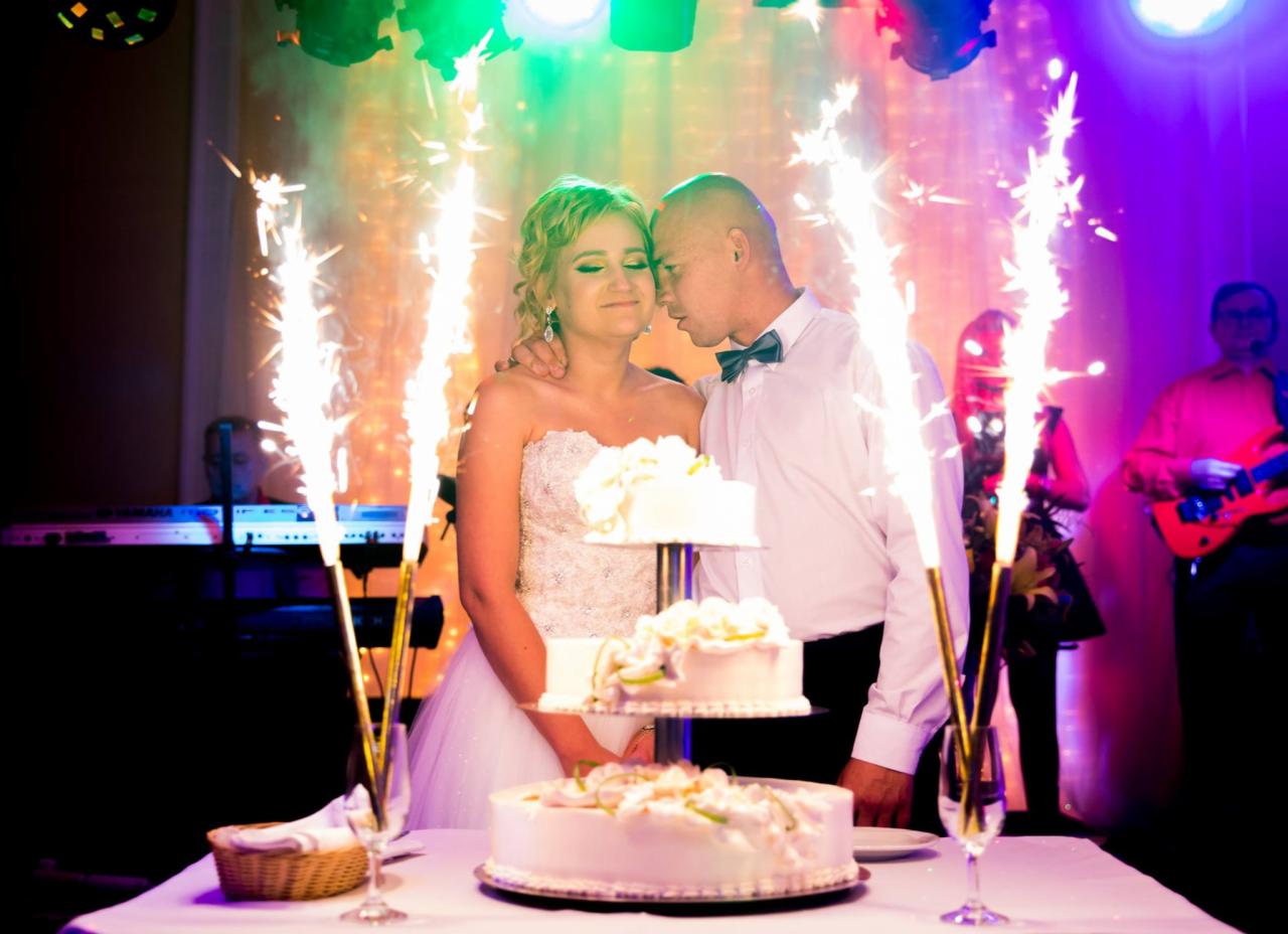 Para młoda stoi przy trzypiętrowym torcie weselnym, otoczona fontannami iskier, w tle zespół muzyczny i kolorowe oświetlenie.