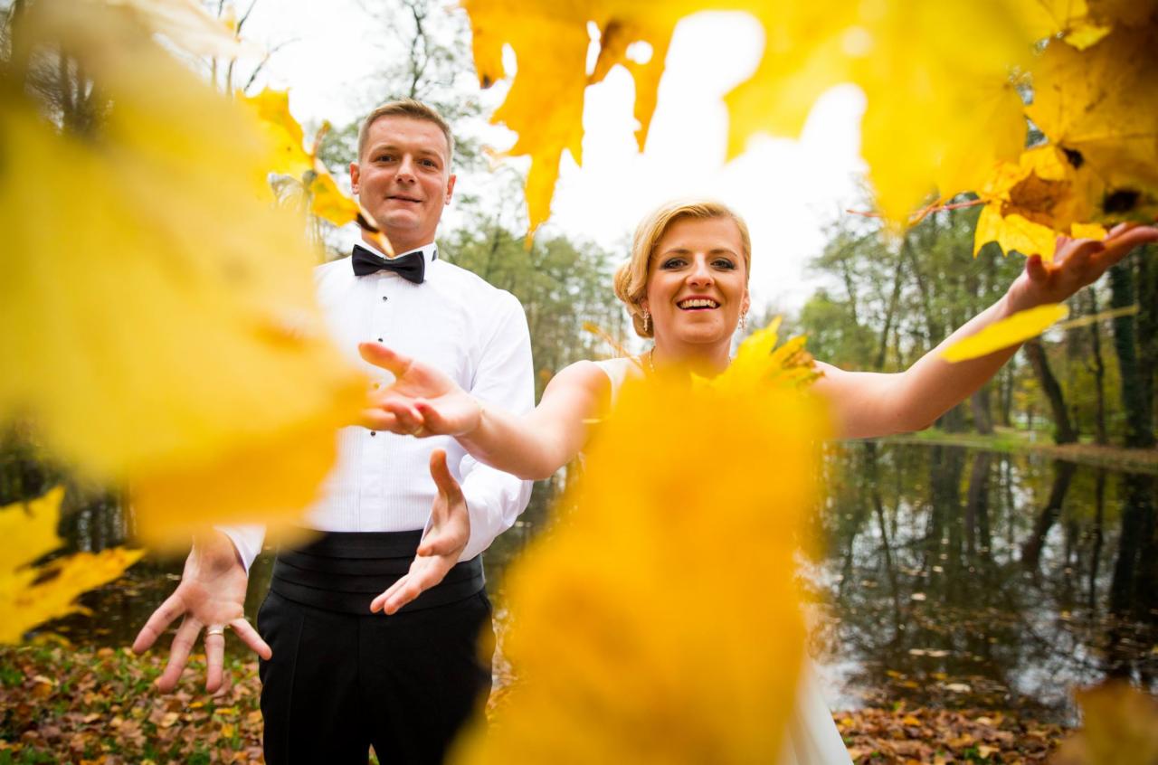 Uśmiechnięta para młoda w jesiennej scenerii rzuca w górę żółte liście; w tle widoczne drzewa i tafla wody.