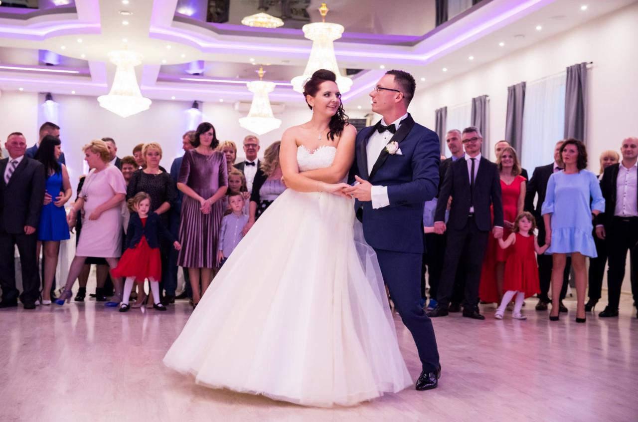 Para młoda w tańcu na sali weselnej, w tle zgromadzeni goście w różnorodnych strojach, oświetlenie sufitowe z ozdobnymi żyrandolami.