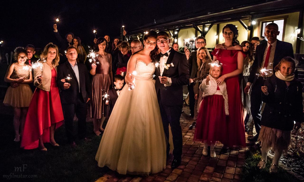 Para młoda z gośćmi weselnymi świętuje nocą, trzymając zapalone zimne ognie na tle budynku.