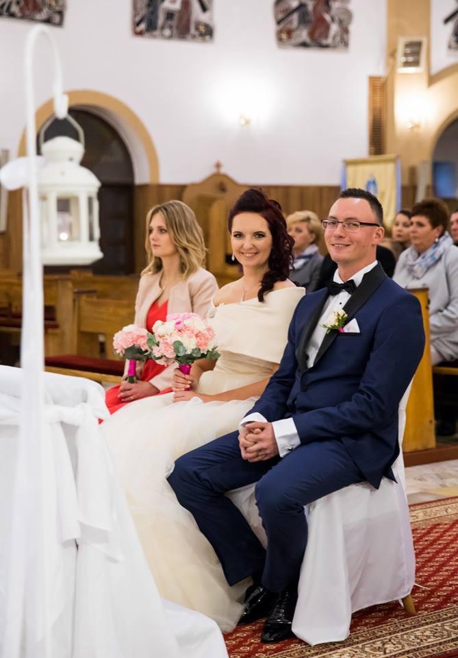 Para młoda siedzi podczas ceremonii w kościele. Panna młoda w sukni z odkrytymi ramionami trzyma bukiet różowych i białych kwiatów. Pan młody w granatowym smokingu z czarną muchą i butonierką.