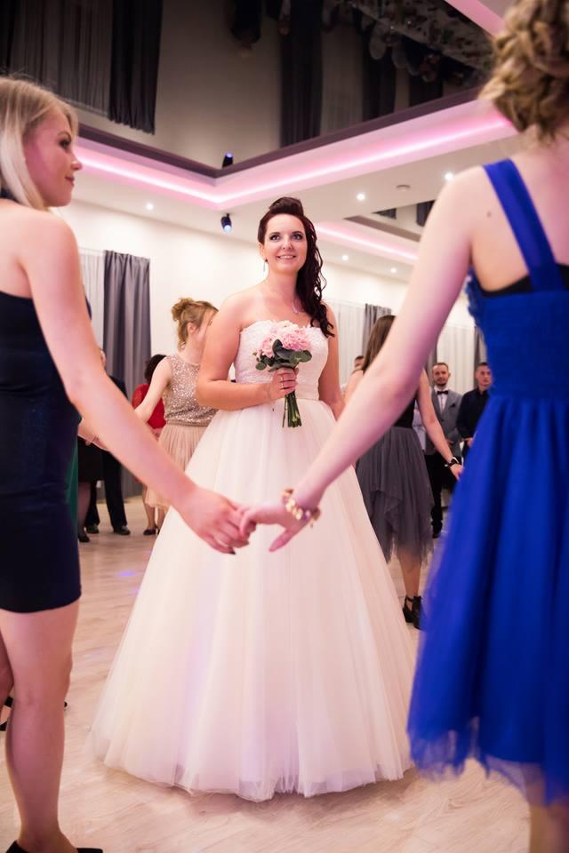 Panna młoda w białej sukni trzymająca bukiet różowych kwiatów, otoczona przez druhny podczas tradycyjnego tańca w kręgu na sali weselnej.