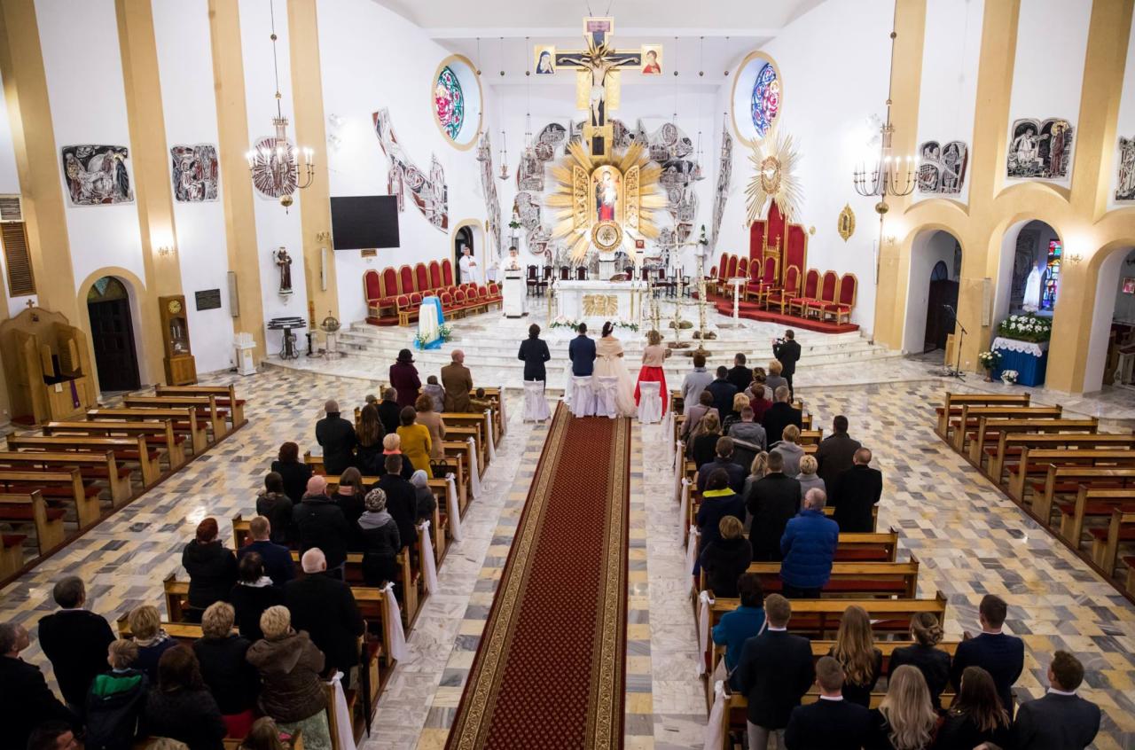 Para młoda stoi przed ołtarzem w kościele, goście siedzą w ławkach po obu stronach czerwonego dywanu, widok z góry.