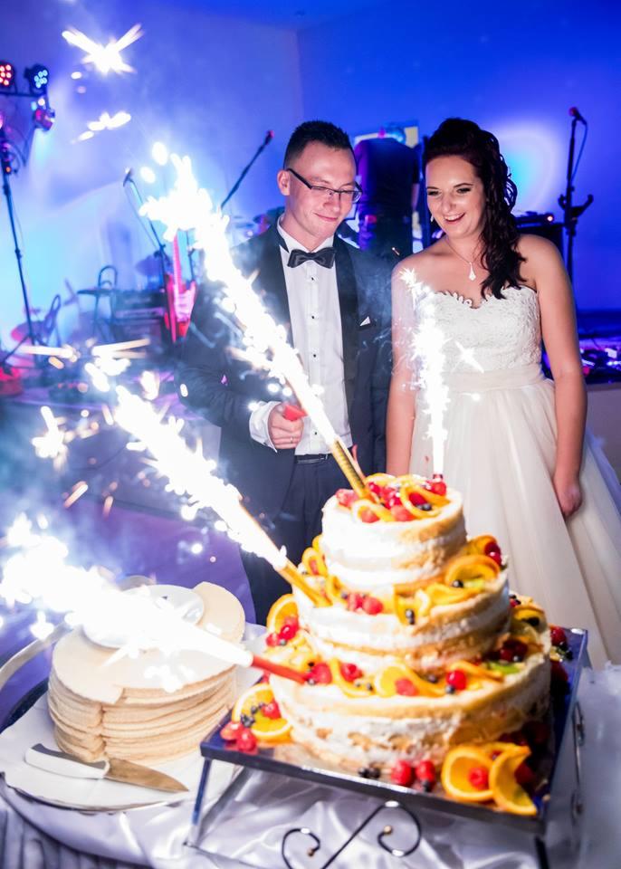 Uśmiechnięta para młoda, w tle scena z instrumentami, kroi tort weselny z owocami i zimnymi ogniami.