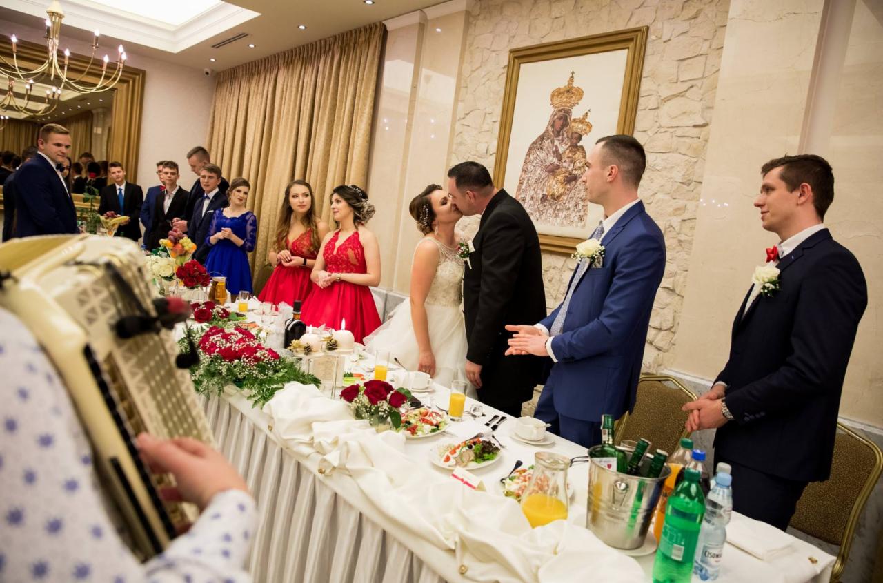 Nowożeńcy całują się za stołem weselnym udekorowanym kwiatami i jedzeniem, w tle goście i obraz Matki Boskiej Częstochowskiej.