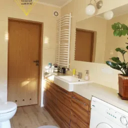 Jasna łazienka z drewnianymi szafkami pod umywalką, pralką i grzejnikiem drabinkowym, okrągły dywanik na podłodze z płytek imitujących drewno.