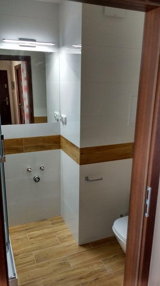 Nowoczesna łazienka z białymi płytkami, drewnopodobną podłogą i akcentem z drewnianych paneli na ścianie, widoczna toaleta i armatura prysznicowa.