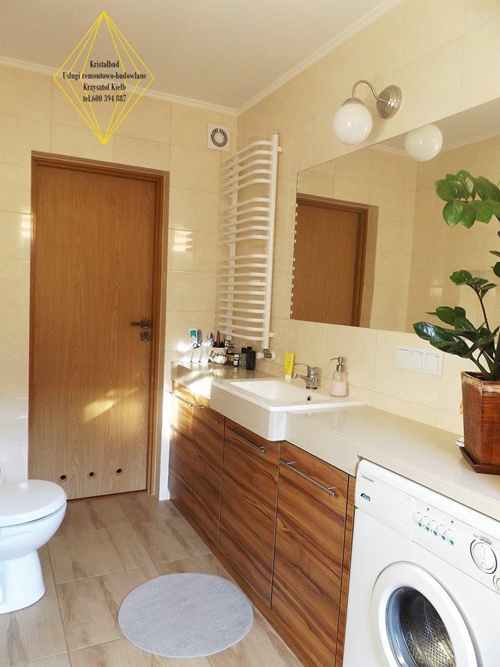 Jasna łazienka z drewnianymi szafkami pod umywalką, pralką i grzejnikiem drabinkowym, okrągły dywanik na podłodze z płytek imitujących drewno.