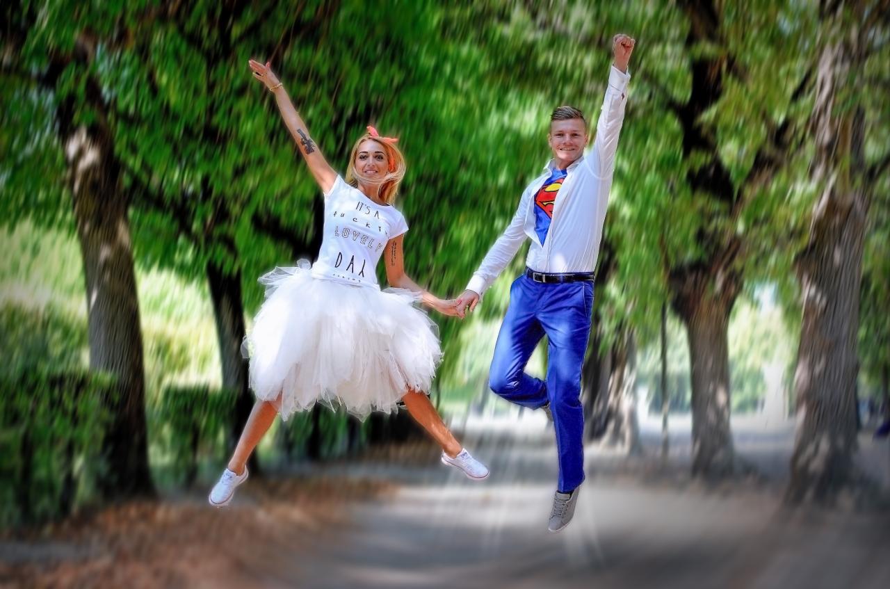 Para młoda w strojach superbohaterów skacze w parku, trzymając się za ręce. Ona w białej tiulowej spódnicy i koszulce z napisem, on w błękitnych spodniach i rozpiętej koszuli z logo Supermana.