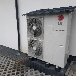 Zewnętrzna jednostka pompy ciepła LG Therma V, zamontowana na betonowych podstawach z daszkiem ochronnym z grafitowej blachodachówki, na tle białej ściany budynku i szarej kostki ażurowej.