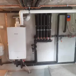 Biały wiszący piec gazowy Junkers z podłączonymi rurami miedzianymi i czarnymi izolowanymi, szare ściany i sufit w surowym stanie, widoczny rozdzielacz ogrzewania podłogowego i sterownik pomarańczowy.