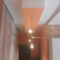 Wnętrze w trakcie prac wykończeniowych: pomarańczowa ściana z białym sufitem, zabezpieczona folią ochronną, widoczne wiszące lampy i fragmenty drewnianej konstrukcji.