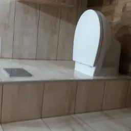 Biała toaleta z deską na podwyższeniu wykończonym płytkami imitującymi drewno, z widocznym odpływem liniowym i półką z środkami czystości.