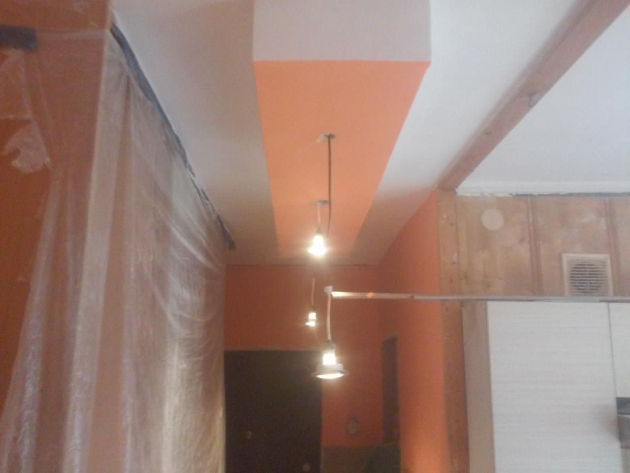 Wnętrze w trakcie prac wykończeniowych: pomarańczowa ściana z białym sufitem, zabezpieczona folią ochronną, widoczne wiszące lampy i fragmenty drewnianej konstrukcji.