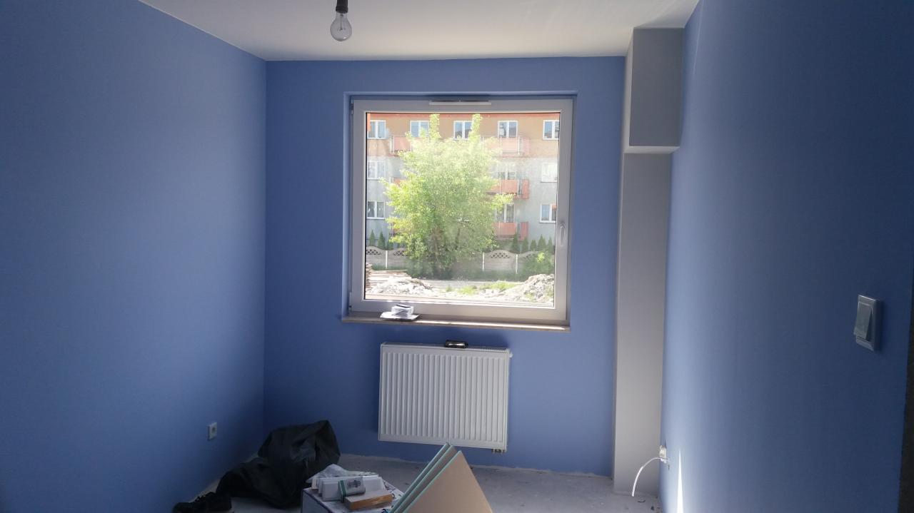 Pokój po odświeżeniu ścian farbą w odcieniu niebieskiego, widoczny grzejnik, okno z widokiem na zieleń i budynek, na podłodze resztki materiałów budowlanych.