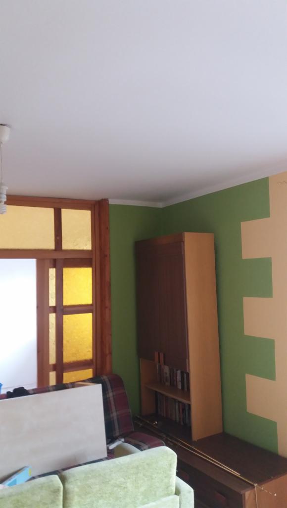 Pokój po odświeżeniu ścian: zielona farba na ścianie z beżowymi, geometrycznymi wzorami, drewniana szafka z książkami, sofa w tle.