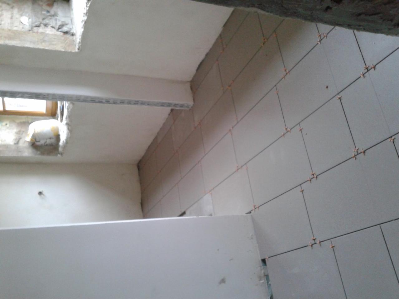 Świeżo ułożone szare płytki ścienne z pomarańczowymi krzyżykami dystansowymi, widoczne fragmenty surowego stropu i ściany z oknem w tle.