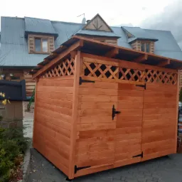 Drewniana szopa ogrodowa z dwuskrzydłowymi drzwiami i kratownicą u góry, na tle drewnianego domu z blachodachówką i workami na palecie.