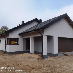 Nowoczesny dom jednorodzinny z jasną elewacją, ciemną dachówką i brązową bramą garażową, widoczny z zewnątrz na działce w trakcie zagospodarowania.