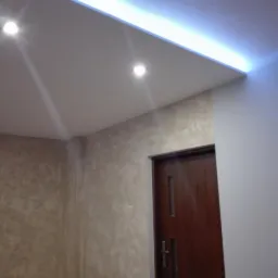 Nowoczesne wnętrze z oświetleniem LED w suficie podwieszanym, dekoracyjna ściana w odcieniach beżu i brązu oraz gładka, pomalowana na biało ściana obok ciemnych drzwi.