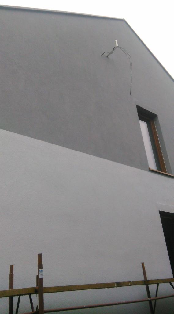 Ściana budynku z częściowo wykonanym tynkiem w dwóch odcieniach szarości, widoczne rusztowanie i wystające kable.