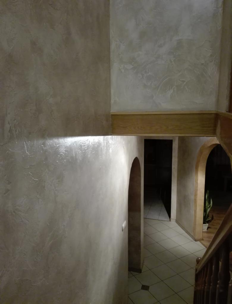 Korytarz z dekoracyjną ścianą w odcieniach beżu, z wyraźną fakturą i delikatnym połyskiem, wykończony drewnianym elementem architektonicznym, widoczne łuki prowadzące do dalszej części domu.