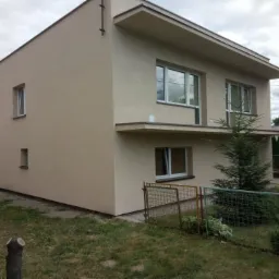 Dwupiętrowy dom jednorodzinny po remoncie elewacji w odcieniach beżu, z widoczną zielenią w ogrodzie i fragmentem starego budynku w tle.