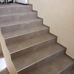 Schody wyłożone brązowymi płytkami ceramicznymi z metalowymi listwami na krawędziach, widok z dołu na górę, ściana w kolorze ecru z fakturą.