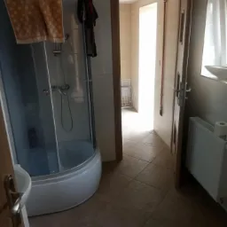 Łazienka z kabiną prysznicową z brązowym ręcznikiem na górze, brązowymi płytkami na podłodze, białym grzejnikiem i otwartymi drzwiami do kolejnej łazienki, widok z perspektywy wejścia.