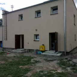 Dwukondygnacyjny budynek o beżowej elewacji z dwiema otwartymi, ciemnobrązowymi drzwiami wejściowymi, zewnętrznymi schodami betonowymi, drabina stalowa na ścianie, żółta skrzynka gazowa i widoczne...