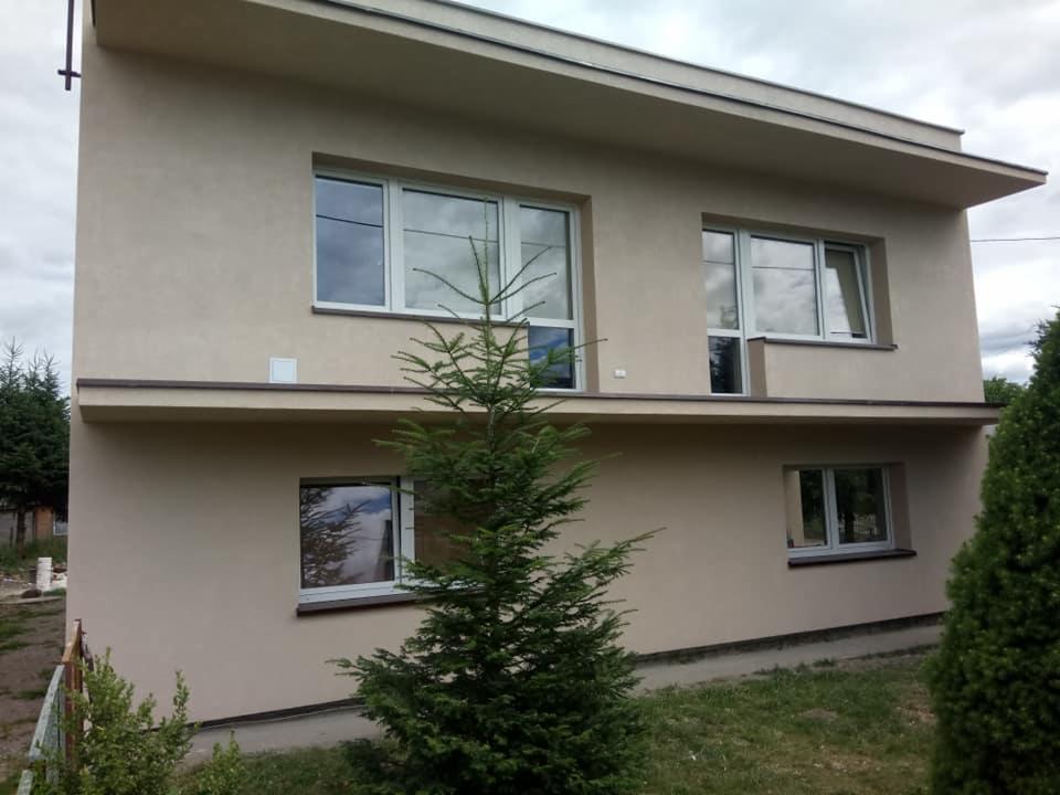 Dwukondygnacyjny dom z beżową elewacją, białymi ramami okiennymi i wystającym dachem, widziany z poziomu gruntu przez iglaste drzewo.