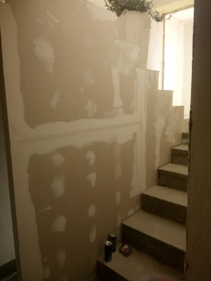 Ściana połatana gipsem i schody w trakcie remontu w Trzebiatowie, widoczne puszki po napojach i paczka papierosów na schodach.