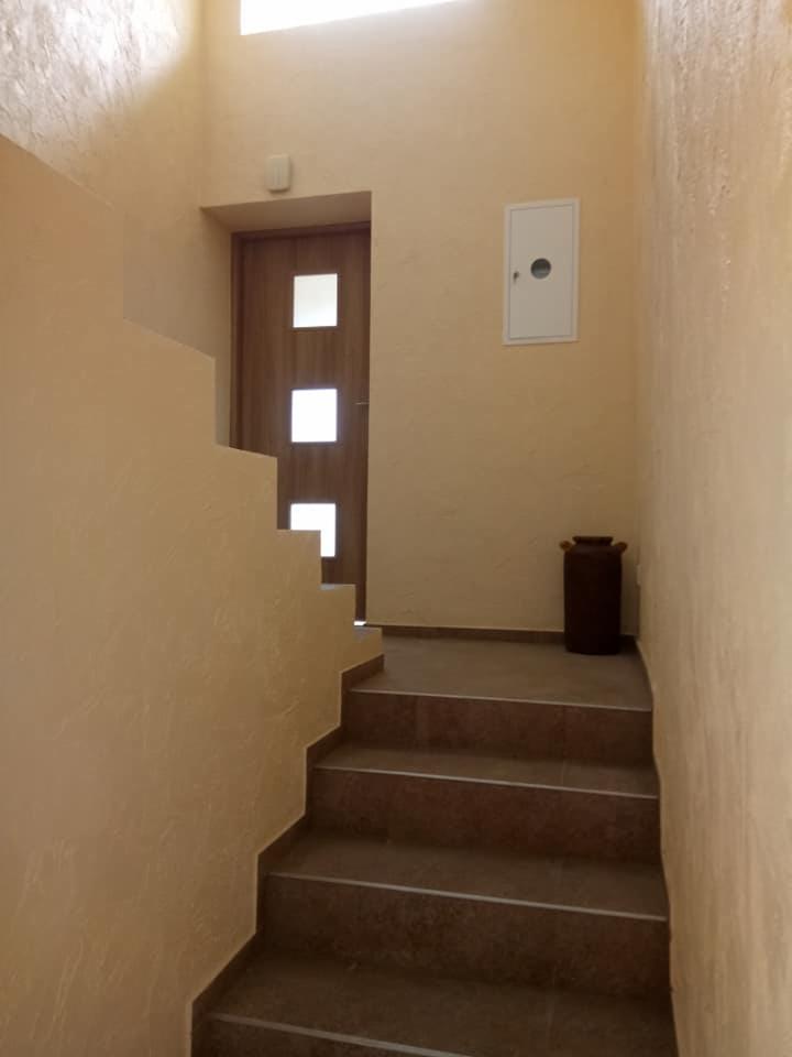 Wnętrze klatki schodowej z brązowymi schodami wyłożonymi płytkami, drewnianymi drzwiami z trzema kwadratowymi przeszkleniami i beżowymi ścianami o wyraźnej fakturze.