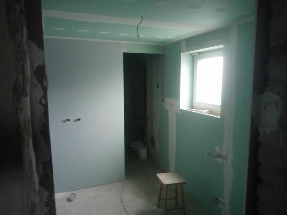 Wnętrze łazienki w trakcie remontu z płytami gipsowo-kartonowymi na ścianach, widoczne otwory na instalacje, białe fugi i drewniany stołek.