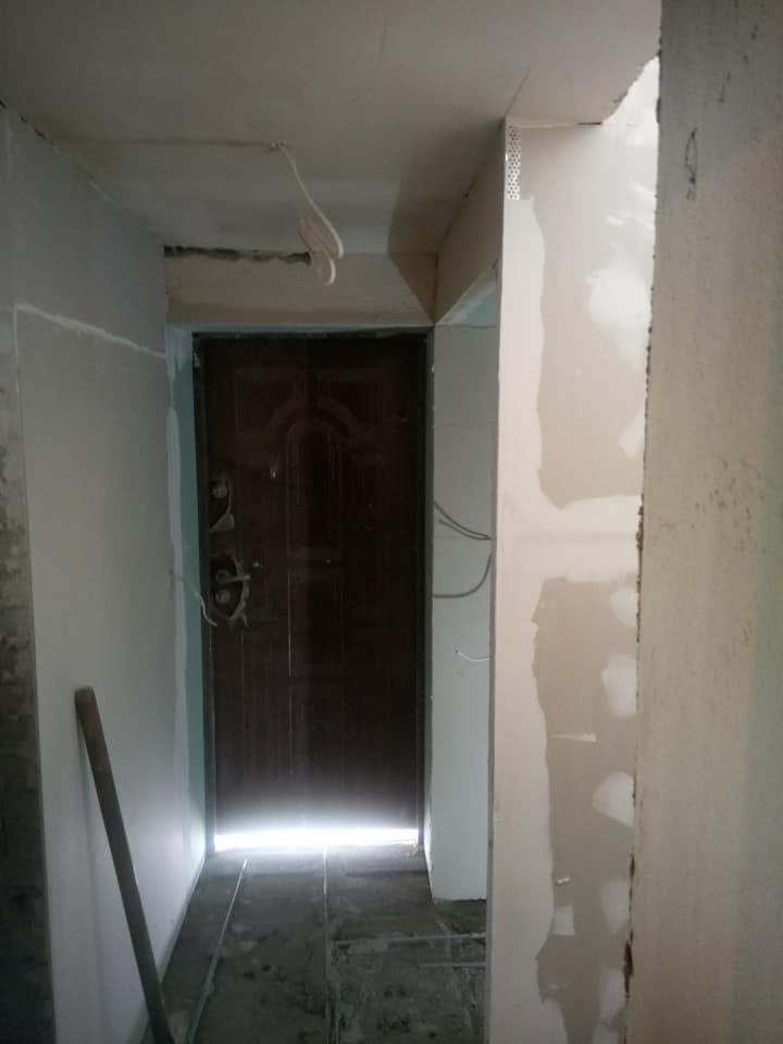 Korytarz w trakcie remontu: ściany częściowo pokryte gładzią, widoczna metalowa siatka na rogu ściany, ciemne drzwi wejściowe z widocznym światłem z zewnątrz, na podłodze widoczne ślady prac...