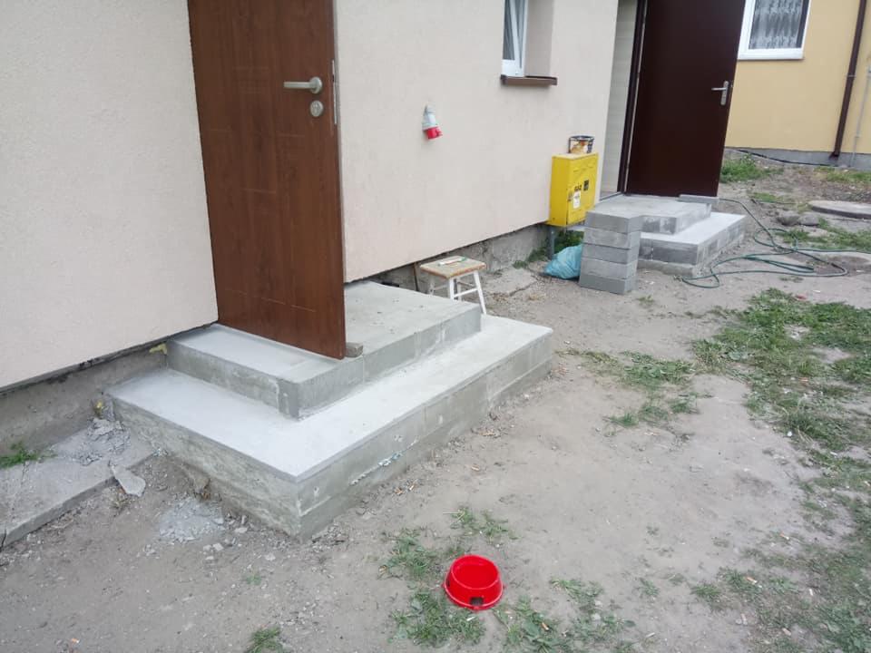 Betonowe schody wejściowe do budynku w trakcie budowy, z niedokończonymi fugami i elementami konstrukcyjnymi, widoczne dwie pary schodów, jedna z brązowymi drzwiami, druga z otwartymi brązowymi...