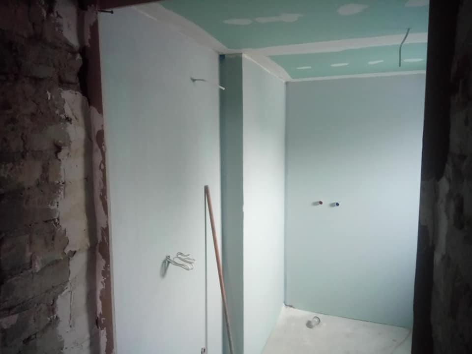 Wnętrze łazienki w trakcie remontu, widoczne surowe ściany z cegły oraz fragment ściany pokrytej płytą gipsowo-kartonową pomalowaną na jasny kolor, wystające kable.