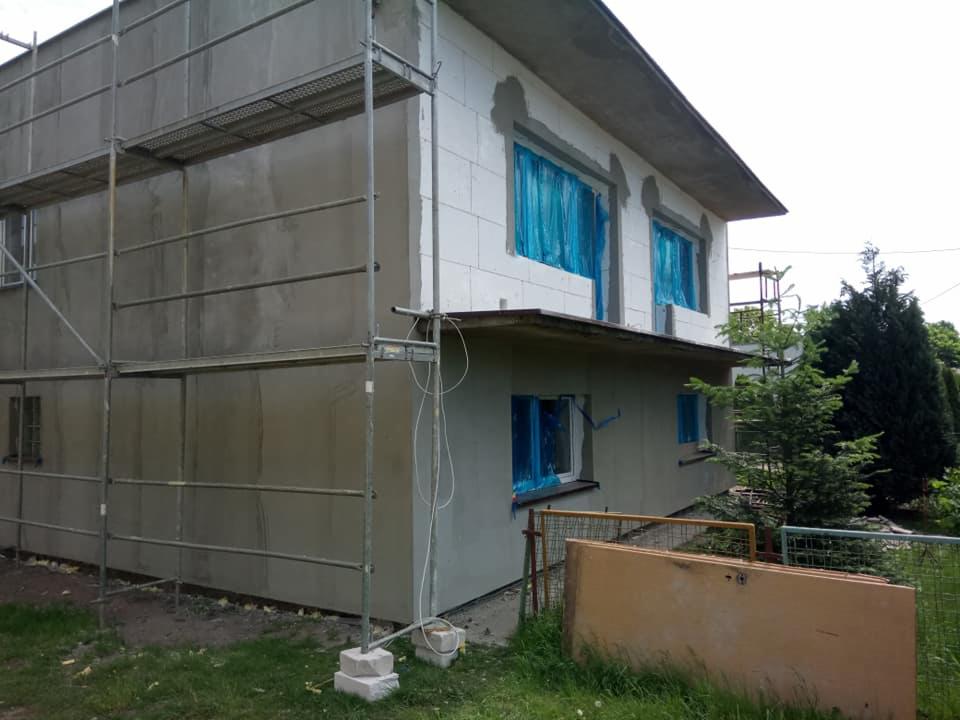 Dom w trakcie ocieplania styropianem, okna zabezpieczone folią, widoczne rusztowania i fragment elewacji pokrytej szarym tynkiem.