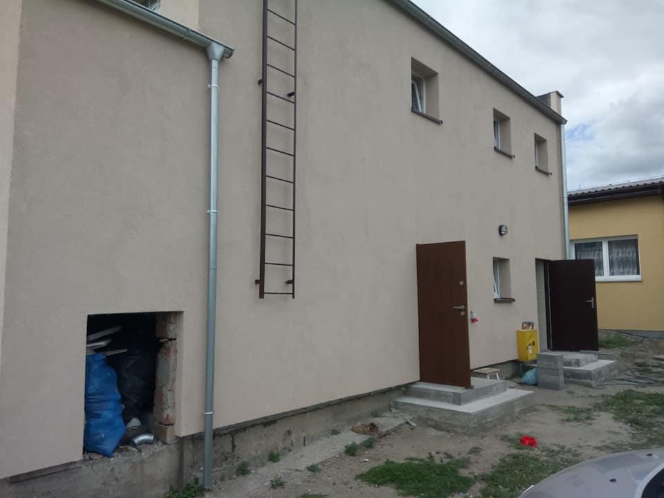 Elewacja budynku po remoncie z widoczną nową elewacją w odcieniach beżu, rynną spustową, drabiną przymocowaną do ściany, dwoma nowymi brązowymi drzwiami wejściowymi z betonowymi schodami...