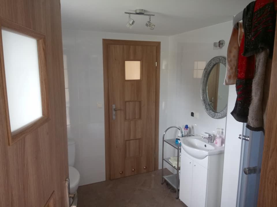 Ukończona łazienka z białymi płytkami, drewnianymi drzwiami z szybą i okrągłym lustrem w srebrnej ramie nad białą szafką z umywalką, widok z otwartych drewnianych drzwi.