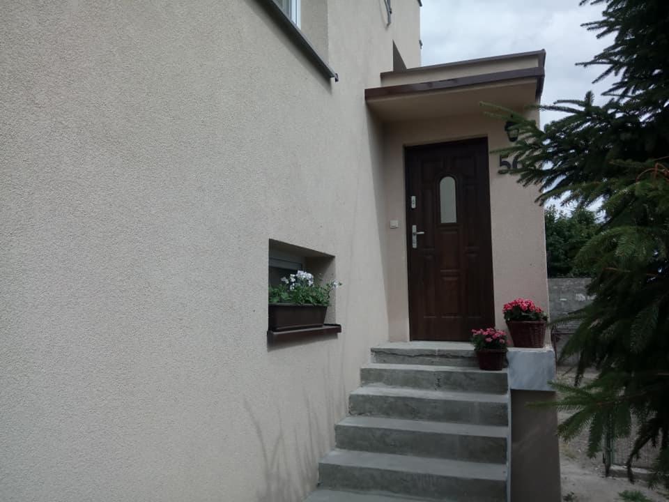 Wejście do domu z drewnianymi drzwiami, betonowymi schodami i oknem z kwiatami, widoczne z boku, numer domu 56.