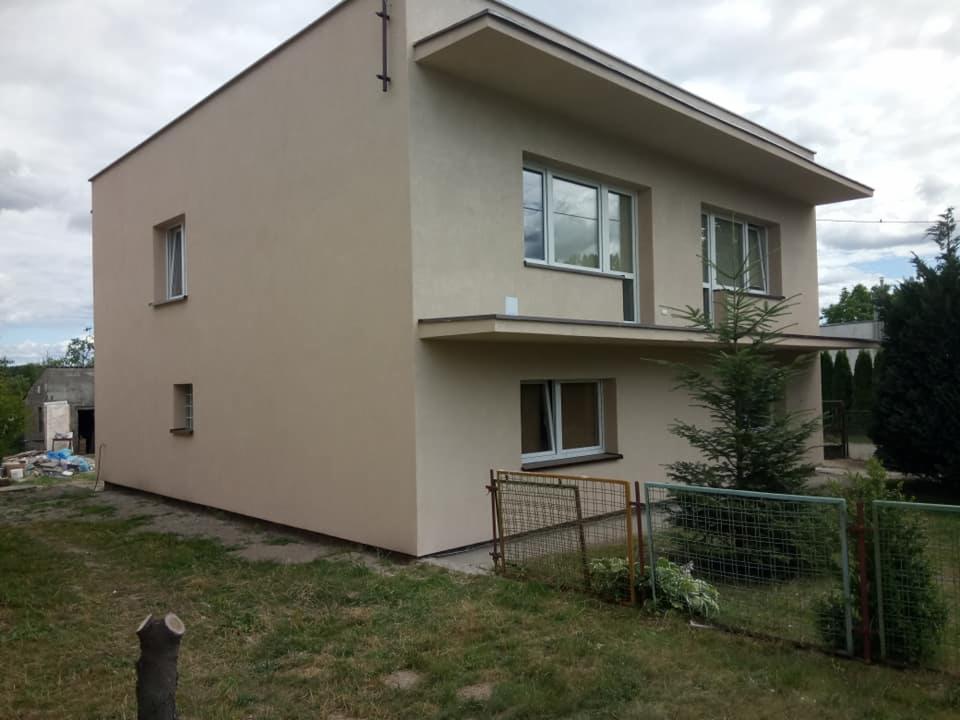 Dwupiętrowy dom jednorodzinny po remoncie elewacji w odcieniach beżu, z widoczną zielenią w ogrodzie i fragmentem starego budynku w tle.