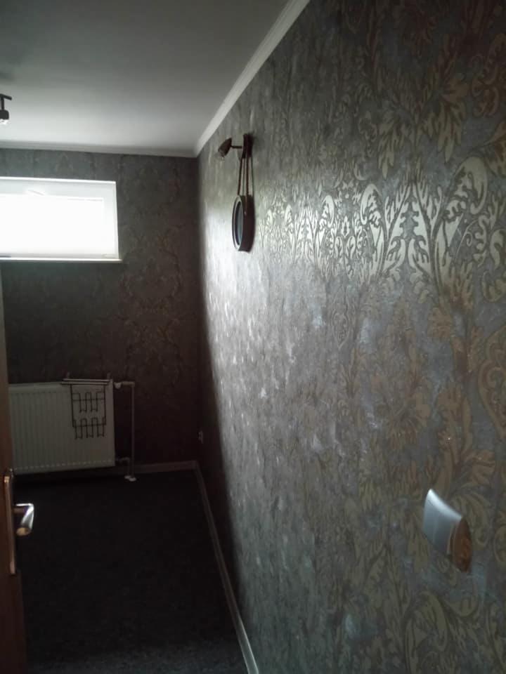 Ściana w korytarzu pokryta dekoracyjną tapetą w odcieniach brązu i złota z motywem roślinnym, widoczny zegar na ścianie i grzejnik w tle.