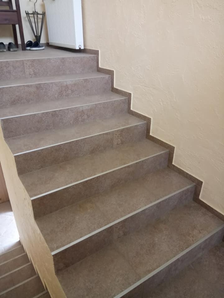 Schody wyłożone brązowymi płytkami ceramicznymi z metalowymi listwami na krawędziach, widok z dołu na górę, ściana w kolorze ecru z fakturą.