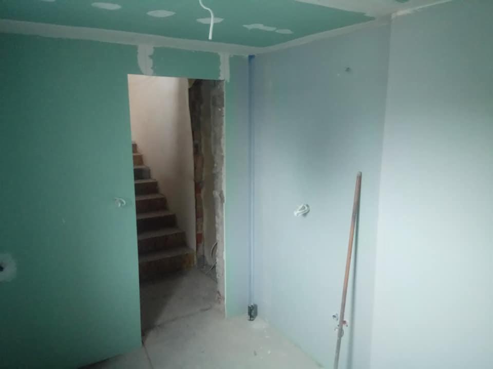 Wnętrze w trakcie prac wykończeniowych: ściany pokryte zielonymi płytami gipsowo-kartonowymi, widoczne schody w przejściu, częściowo zaszpachlowane łączenia płyt.