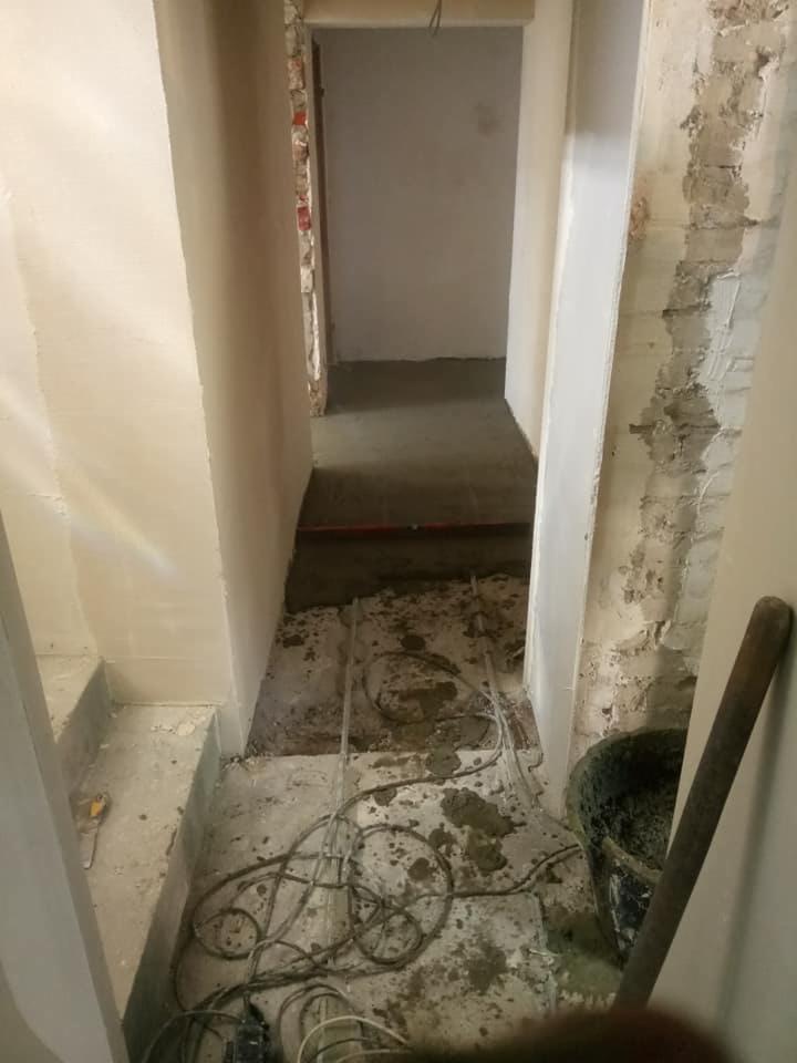 Korytarz w trakcie remontu: ściany częściowo otynkowane, odkryta cegła, widoczne przewody elektryczne, wiadro z zaprawą i mieszadło.