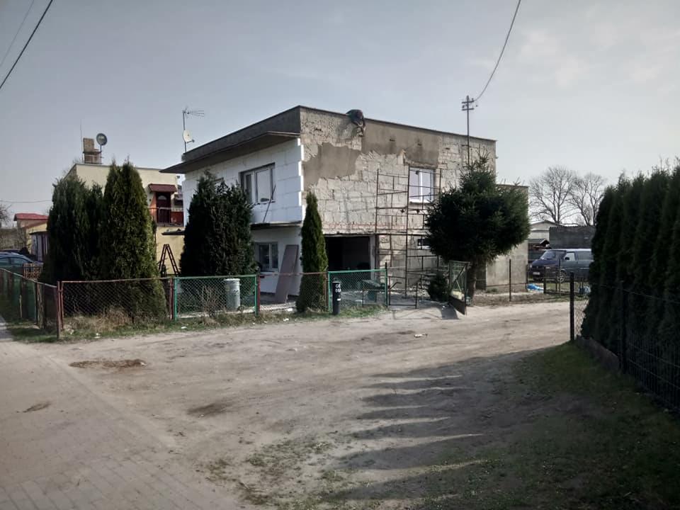 Dwukondygnacyjny dom w trakcie remontu elewacji, widoczne rusztowania, częściowo ocieplona ściana, nierówny teren przed budynkiem.